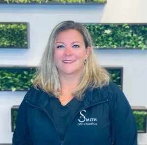Smith Orthodontics team member Sara smiling in office with green plant wall backdrop.