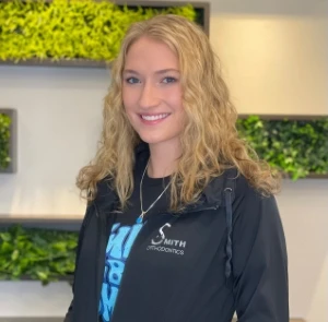 Smith Orthodontics team member Sarah with curly blonde hair smiling in office with green plant wall